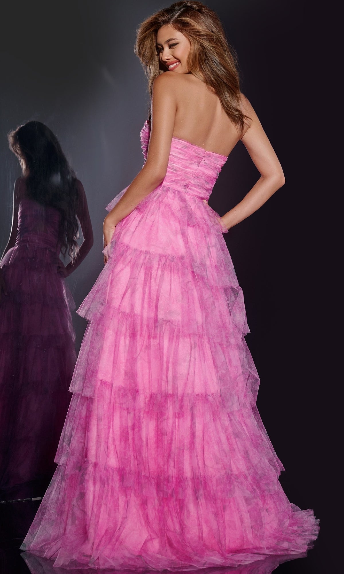 A woman in the Long Prom Dress: Jovani 43170, a gorgeous tiered pink tulle gown, stands before a dark background with her reflection visible behind her.