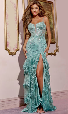 A woman wears the Jovani 43171 Ruffled Long Glitter Prom Dress in sage with sequins, spaghetti straps, a deep neckline, ruffled skirt, and high slit, standing indoors against a decorated wall and framed art.