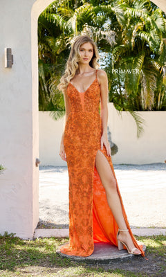 A woman poses near a white archway with greenery, wearing the Primavera 4317 Lace-Up Long Lace Prom Dress in bright orange, featuring embroidered lace, spaghetti straps, and a thigh-high slit.