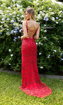 A woman wearing the Primavera 4317 Lace-Up Long Lace Prom Dress in red stands on grass before a bush with white flowers.