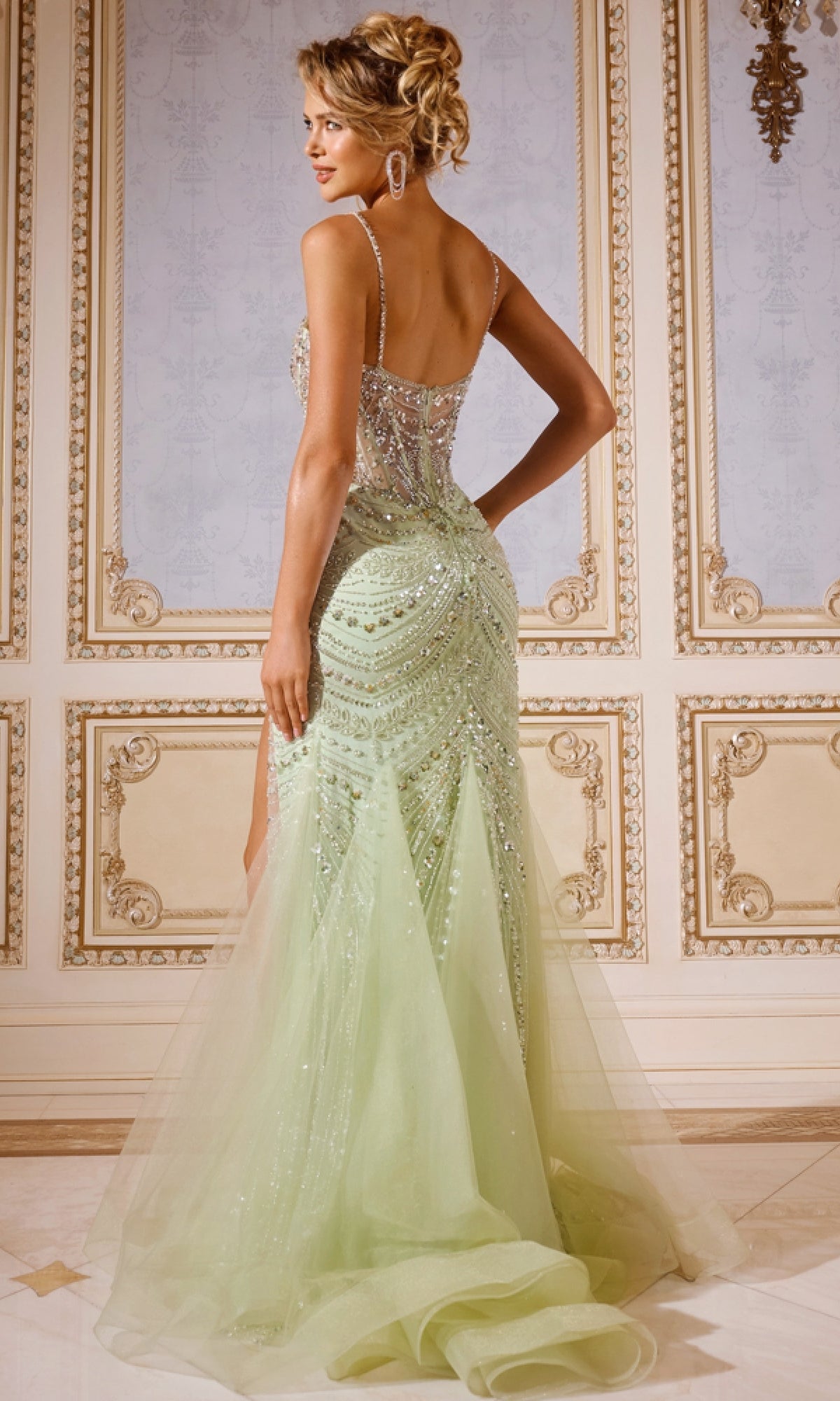 A woman stands with her back to the camera in an ornate room, wearing the Jovani 43189 long prom dress—light green, floor-length mermaid style with a sheer corset bodice, sparkly embellishments, and a tulle train.