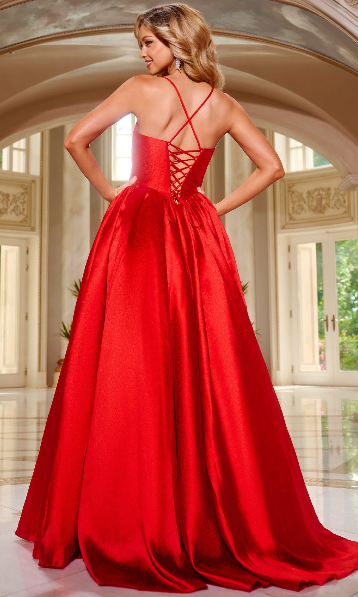 A woman stands in a bright, elegant room wearing the Jovani 43199 Satin Prom Ball Gown—a floor-length red gown with crisscross straps, lace-up back, and corset bodice.