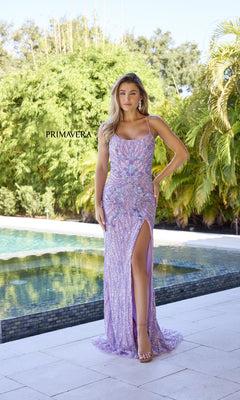 A woman in the Long Beaded Corset Prom Dress: Primavera 4319, a sparkly lavender gown with a high slit, stands by a pool amidst lush greenery. The words "PRIMAVERA" and "Primavera 4319" are shown on the image.