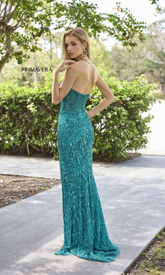 A woman in the Primavera 4321 Strapless Long Sequin Prom Dress with a sweetheart neckline stands on a sidewalk, touching her hair. Green bushes and trees create a lush backdrop for her sparkling sequin evening gown.