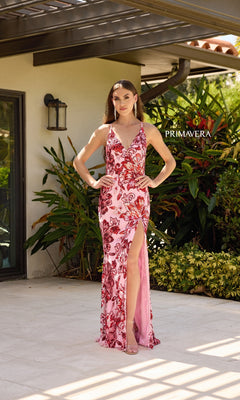 A woman poses confidently outdoors in the Primavera 4323 long prom dress, a sleeveless pink gown with red floral patterns and a v-neckline, set against lush green plants and a modern house.