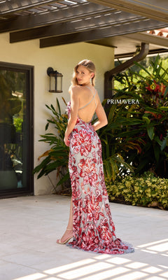 A woman stands outdoors, looking over her shoulder in the Primavera 4323 long prom dress—a backless, floor-length gown adorned with red and white floral sequins. Lush plants and a modern building are visible behind her.