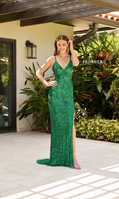 A woman stands on a patio, surrounded by lush plants and a house exterior, wearing the Primavera 4324 long prom dress—a sleeveless, floor-length gown with shimmering sequins and a high side slit.