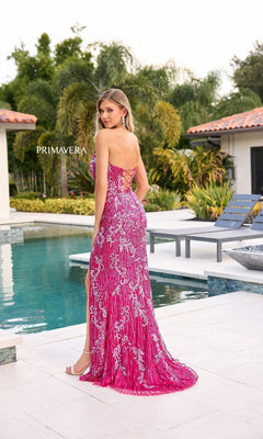 A woman in the Strapless Sequin-Print Prom Gown: Primavera 4328 stands by a pool facing away, with trees and modern buildings creating a striking backdrop for her shimmering formal dress.