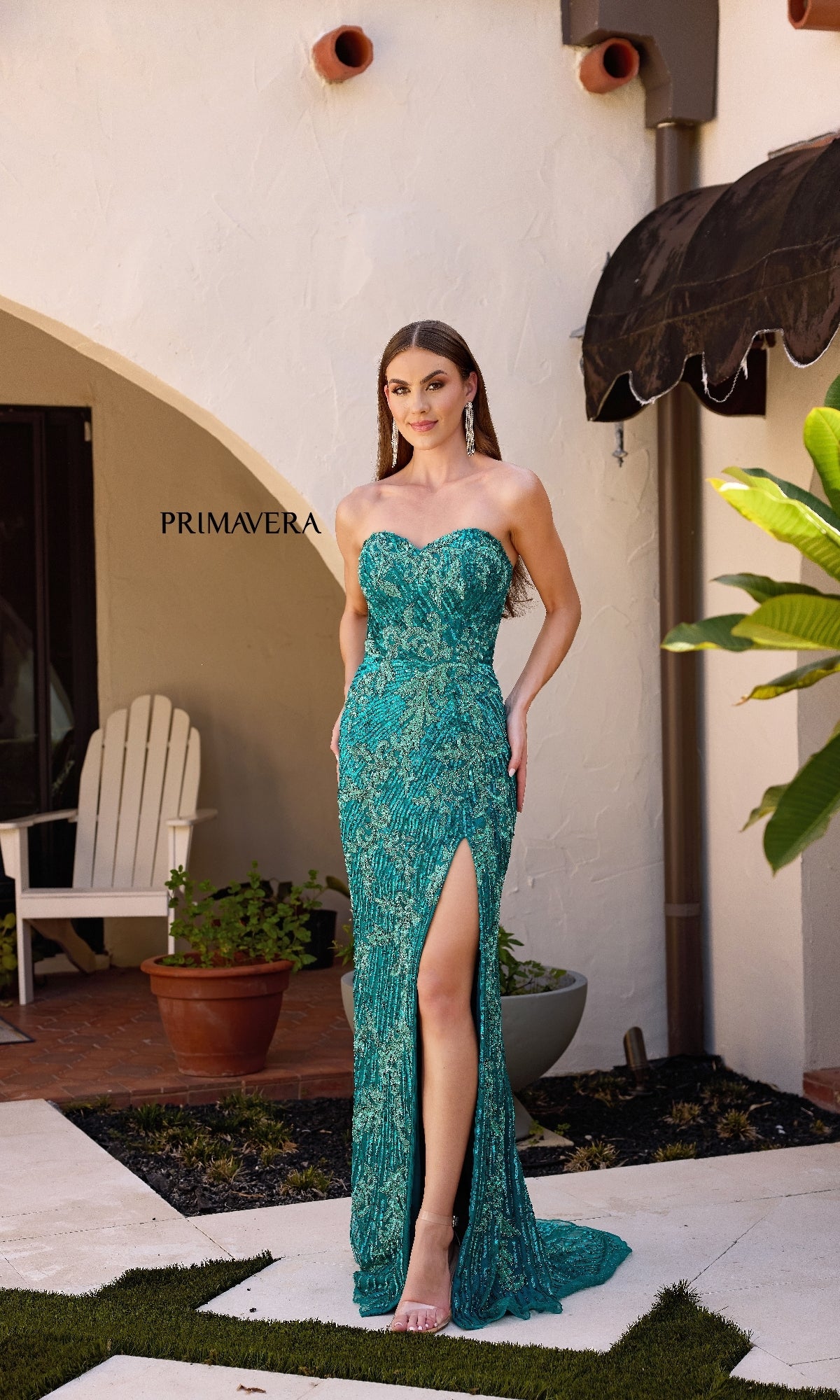 A woman stands by a white stucco building and green plants, elegantly modeling the Strapless Sequin-Print Prom Gown: Primavera 4328 in teal with a high leg slit.