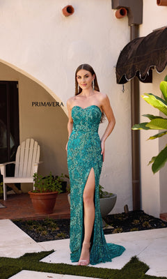 A woman stands by a white stucco building and green plants, elegantly modeling the Strapless Sequin-Print Prom Gown: Primavera 4328 in teal with a high leg slit.