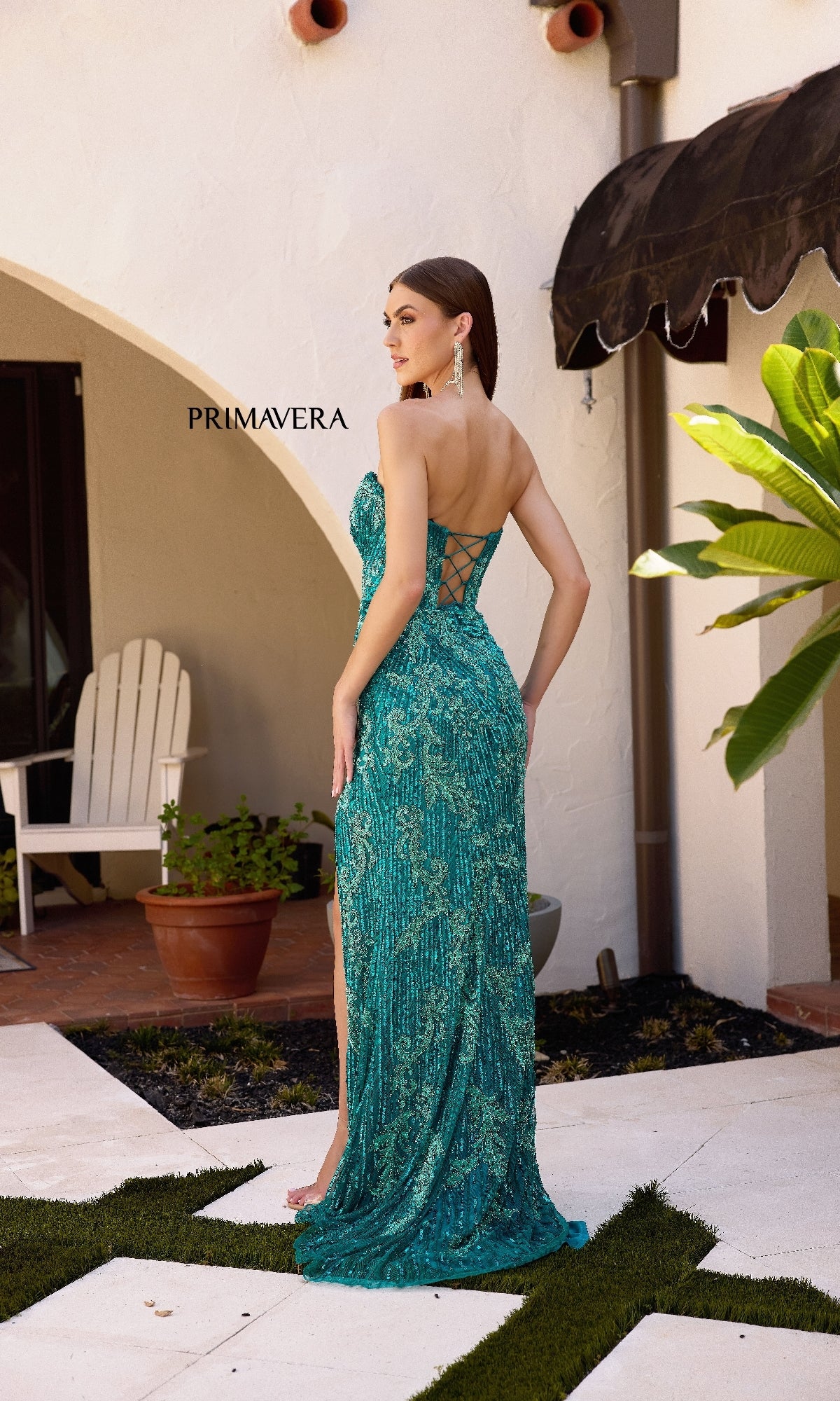 A woman in a Strapless Sequin-Print Prom Gown: Primavera 4328 with a lace-up back and high slit stands outdoors near a white wall and patio furniture.