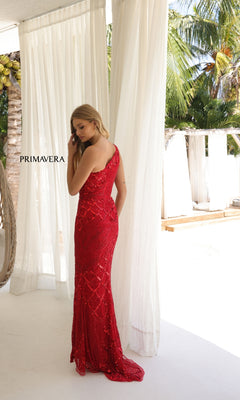 A woman in a red, floor-length sequin gown—Long Prom Dress: Primavera 4329—stands indoors by sheer curtains with palm trees outside. "PRIMAVERA" is displayed on the left curtain as her one-shoulder dress shimmers in the light.