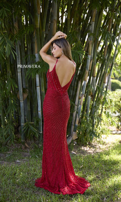 Wearing the Primavera 4330 long prom dress, a woman stands on grass before tall bamboo, dressed in sparkling red sequins with one arm raised and her head turned to the side.
