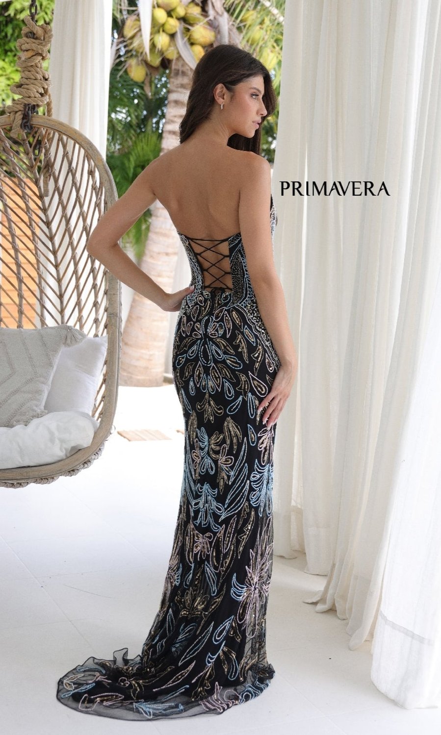 A woman wearing the Strapless Beaded-Print Prom Dress: Primavera 4331, featuring a colorful leaf design, stands by a hanging chair and white curtains with 