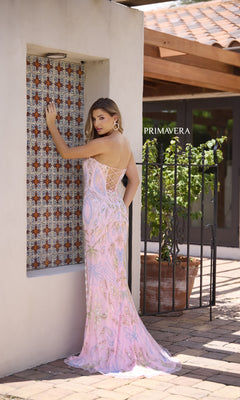 A woman in a Strapless Beaded-Print Prom Dress (Primavera 4331), featuring a lace-up back, stands by a tiled window with a potted plant and wrought iron gate, looking over her shoulder.