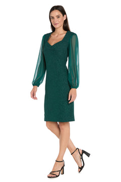 A woman wearing the R&M Richards 4331P petite short green party dress with shimmer fabric and sheer sleeves, paired with black high-heeled sandals, stands against a white background.