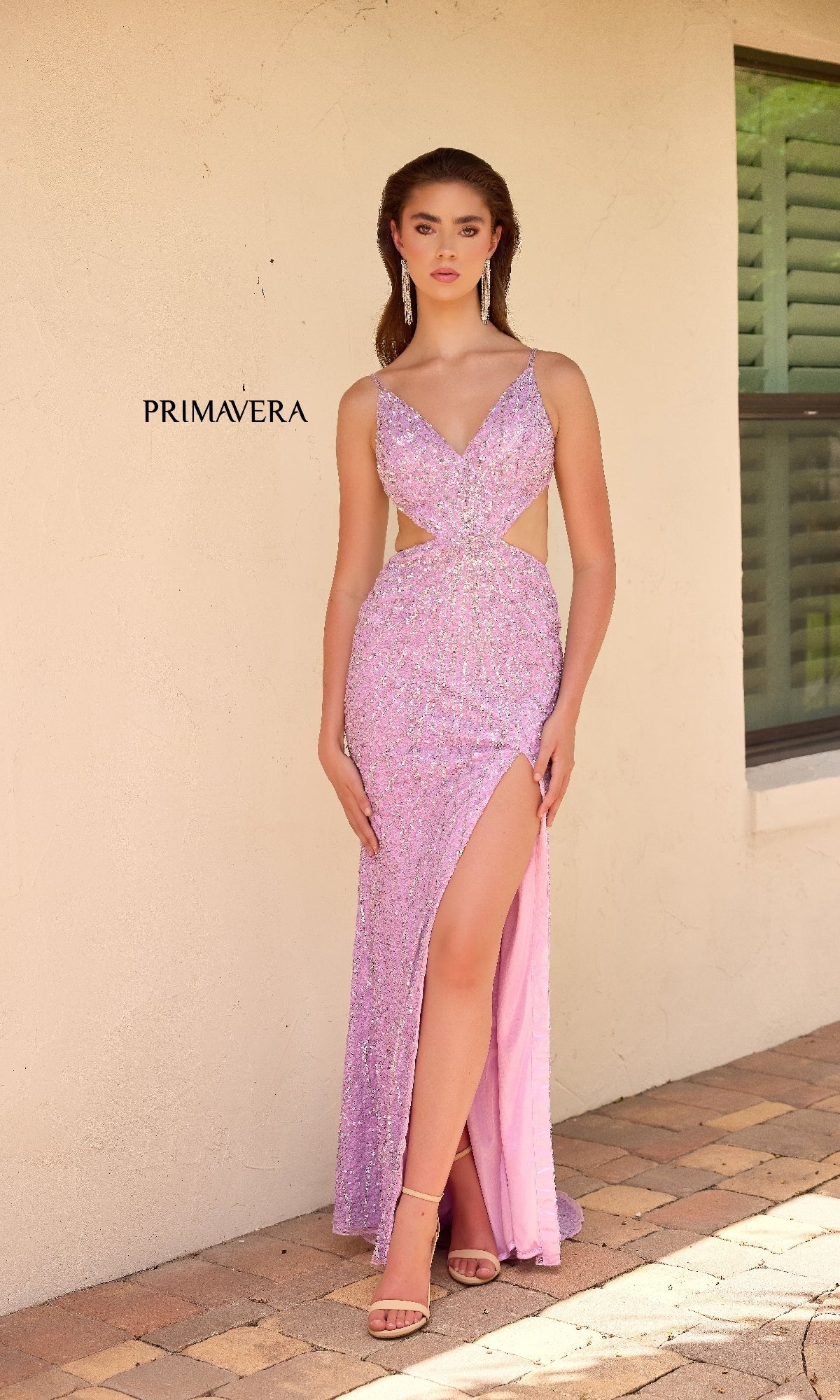 A woman in the Primavera 4334 long prom dress, featuring sparkly light purple sequins, side cut-outs, and a thigh-high slit, stands on a brick patio by a beige wall and window.