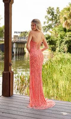 A woman wears the Strapless Long Beaded Prom Dress: Primavera 4335 in coral pink, standing on a wooden deck by a pond with "PRIMAVERA" displayed in the background, facing away from the camera.