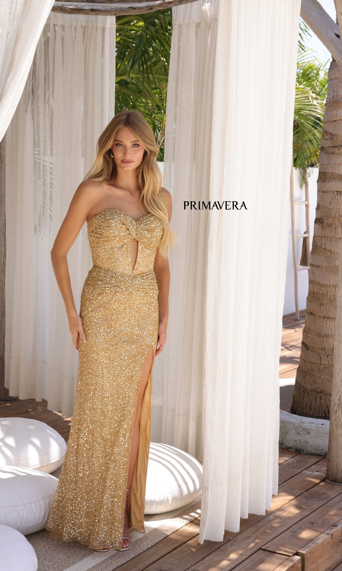A woman wearing the Strapless Long Sequin Prom Dress: Primavera 4340, featuring a gold color and front slit, stands beneath sheer white curtains by a palm tree. 