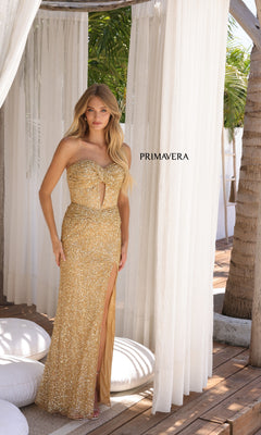 A woman wearing the Strapless Long Sequin Prom Dress: Primavera 4340, featuring a gold color and front slit, stands beneath sheer white curtains by a palm tree. "PRIMAVERA" is visible in the background.