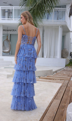 A woman stands outdoors in the Primavera 4341 Long Tiered Sequin Prom Dress, featuring blue ruffled layers and sparkling floral details. A white building and palm fronds enhance the elegant scene.