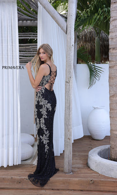 A woman in the Strappy-Back Long Sequin Prom Gown: Primavera 4342, featuring silver floral patterns, stands on a wooden deck by white curtains and a large ceramic vase.