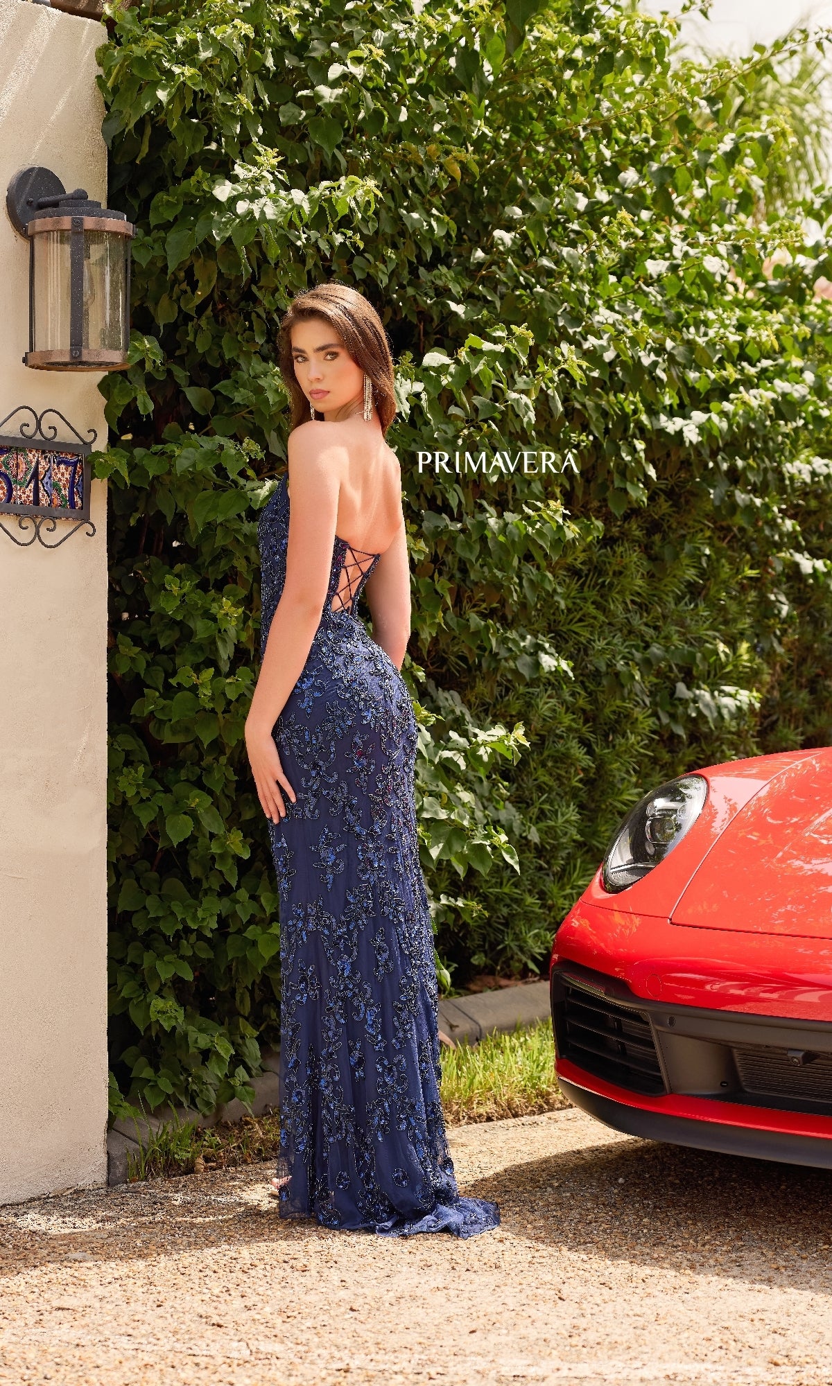A woman in the Strapless Long Sequin Prom Dress: Primavera 4347 stands outdoors by greenery and a red sports car, facing away with her head turned to the camera. 