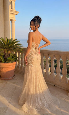 A woman in the Embellished Long Mermaid Formal Prom Dress 4352, featuring a lace-up back, stands on a balcony with sea views. Elegant columns and a potted palm frame her as her shimmering light gold gown catches the sunlight.