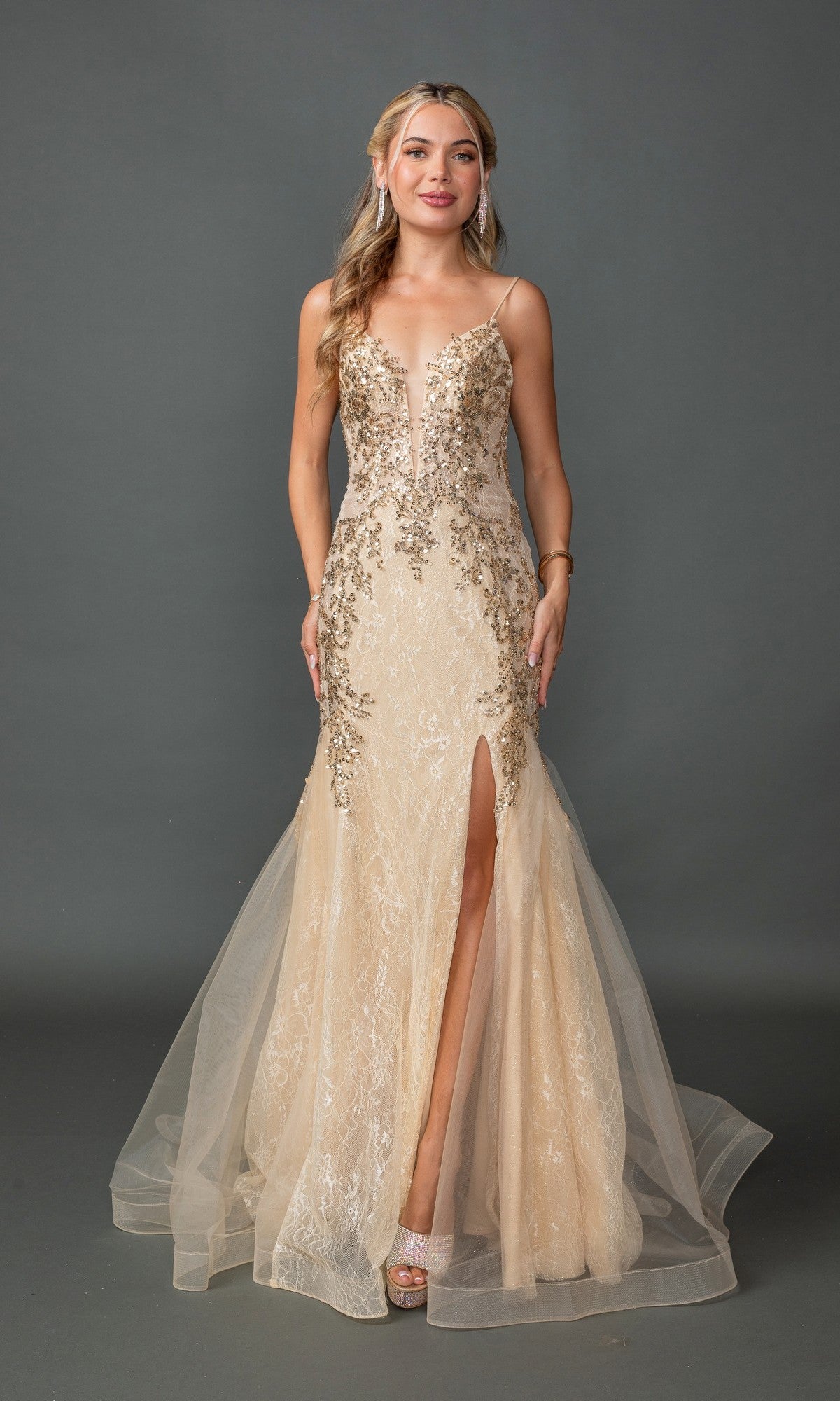 A woman poses against a gray backdrop in the Embellished Long Mermaid Formal Prom Dress 4352 in Champagne, featuring a sleeveless design, deep neckline, high leg slit, and sparkling tulle overlay.