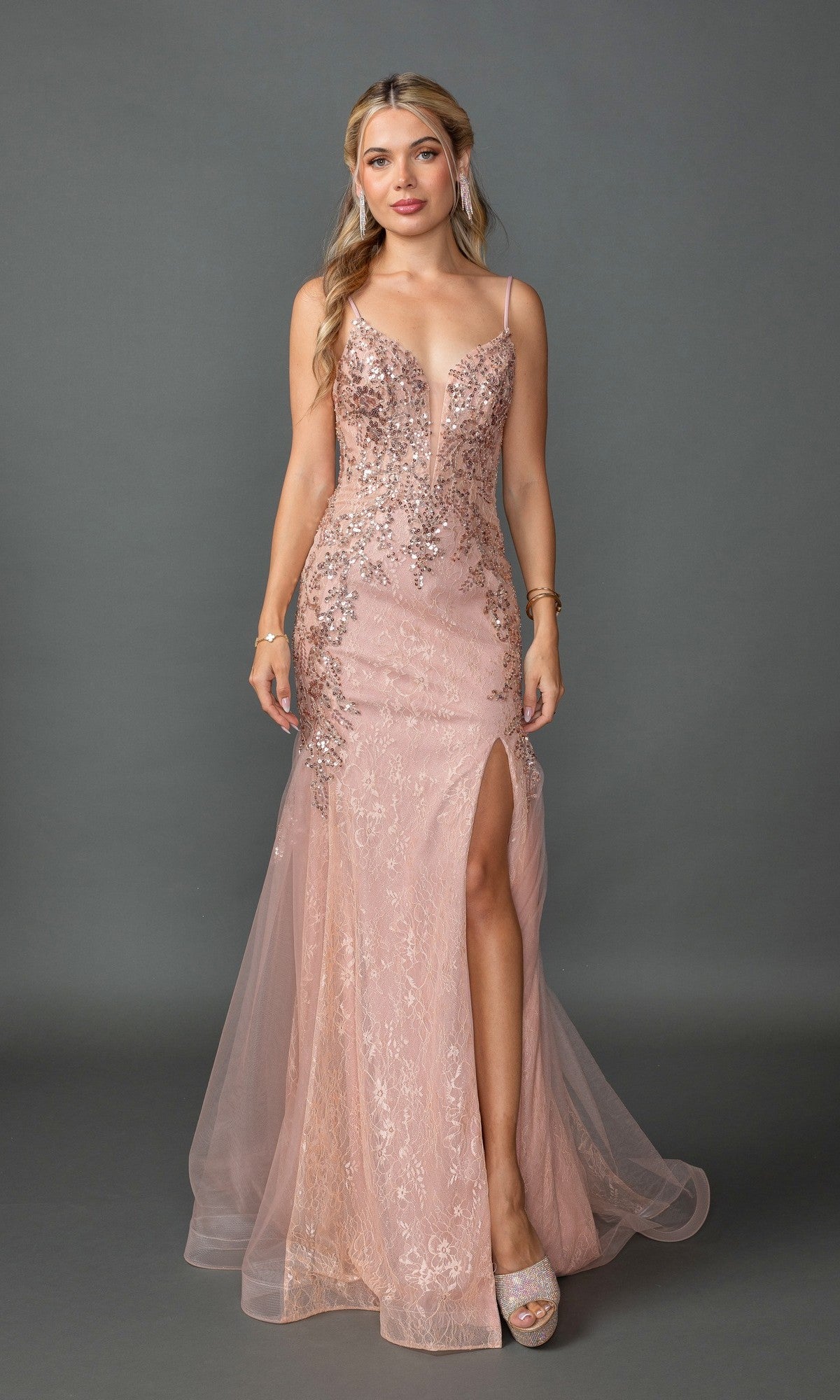 A woman stands against a gray background wearing the Embellished Long Mermaid Formal Prom Dress 4352 in rose gold, featuring a sleeveless design with sequins, deep neckline, high slit, sparkly tulle overlay, and platform heels.