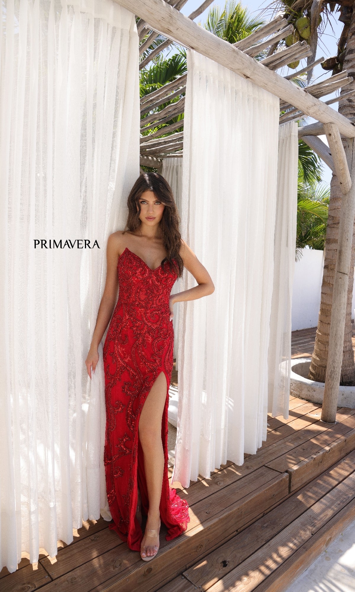 A woman in the Strapless Long Beaded Prom Dress: Primavera 4356, a red, beaded gown with a thigh-high slit, poses on a wooden deck before sheer white curtains featuring the 