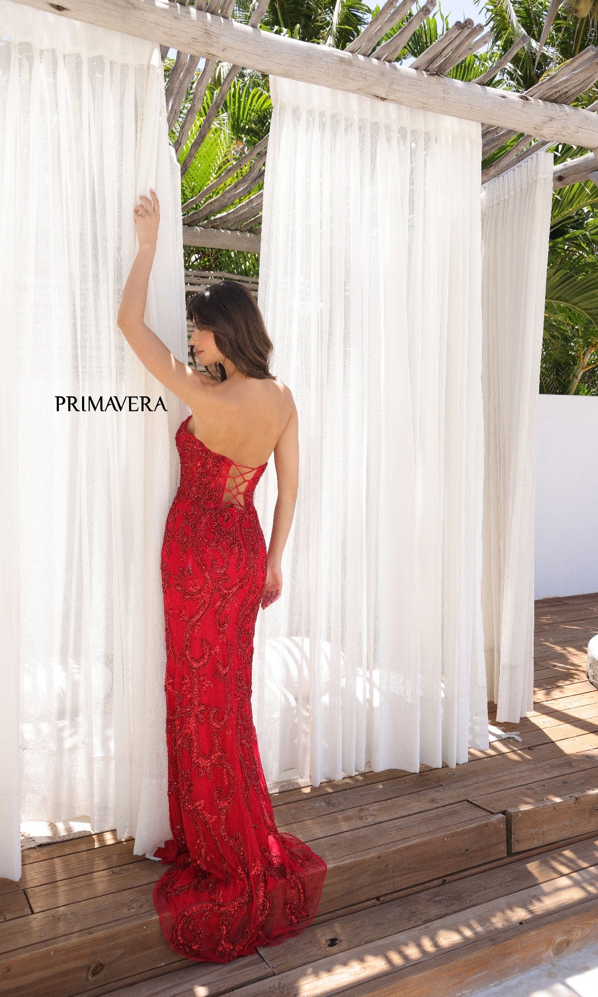 A woman in the strapless Primavera 4356 long beaded prom dress with a lace-up back stands on a wooden deck, facing white sheer curtains with greenery and sunlight behind her.