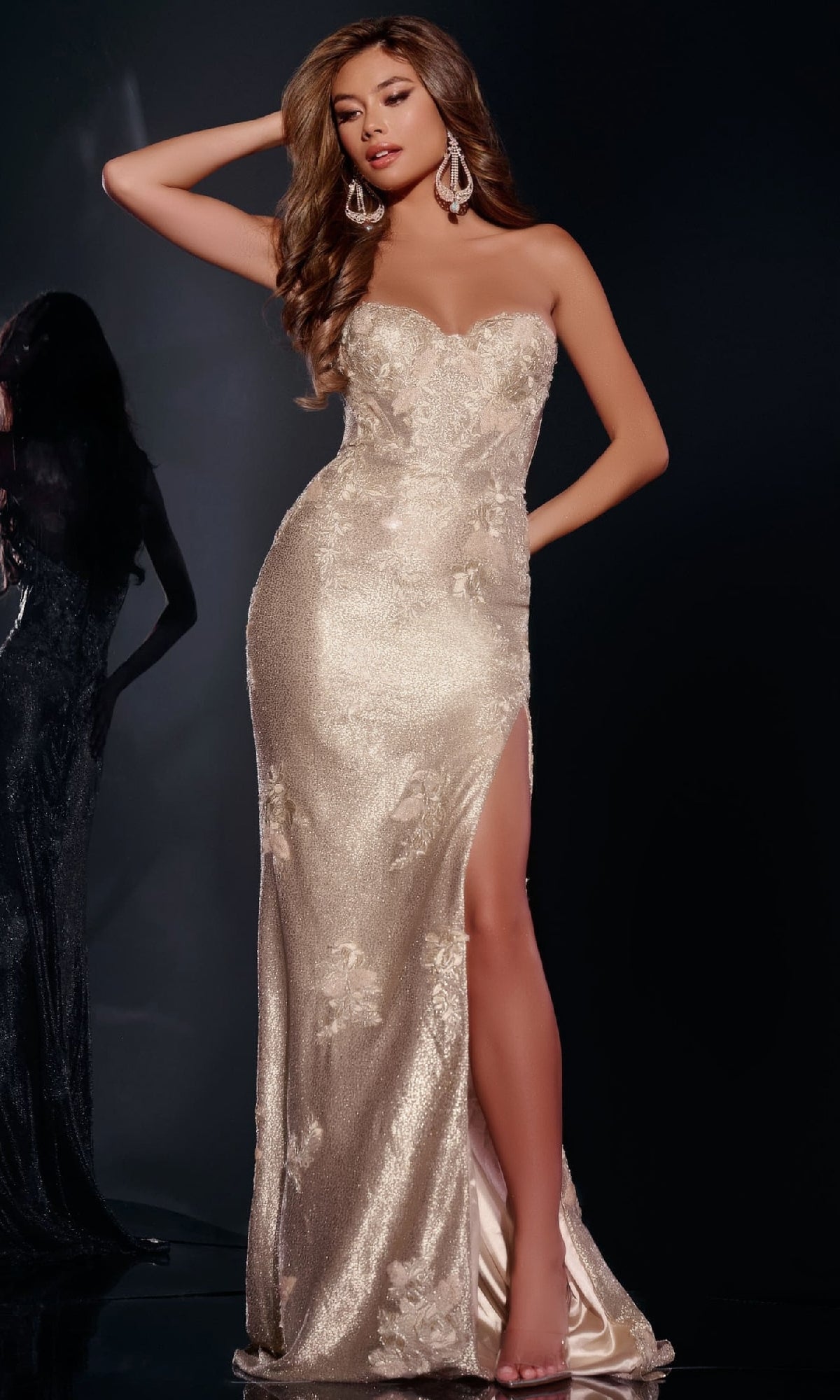 A woman in the Gold Long Prom Dress: Jovani 43587—strapless, fitted gold gown with lace embroidery, thigh-high slit, and floral details—poses with one hand in her hair against a dark background, wearing large earrings.