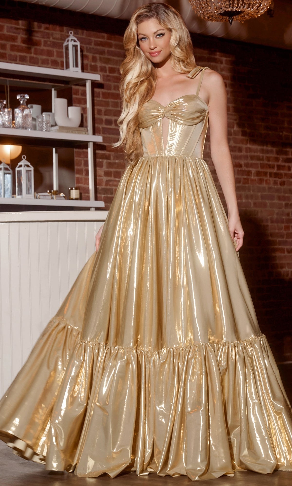 A woman wearing the Jovani 43596 Long Prom Dress, featuring a shiny gold color with a fitted corset bodice and full ruffled skirt, stands indoors near shelves and brick walls.