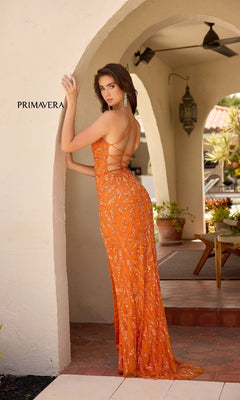 A woman models the Strappy Long Beaded Prom Dress: Primavera 4359 in vibrant orange, posing by a stucco archway outdoors with "PRIMAVERA" displayed on the left wall.