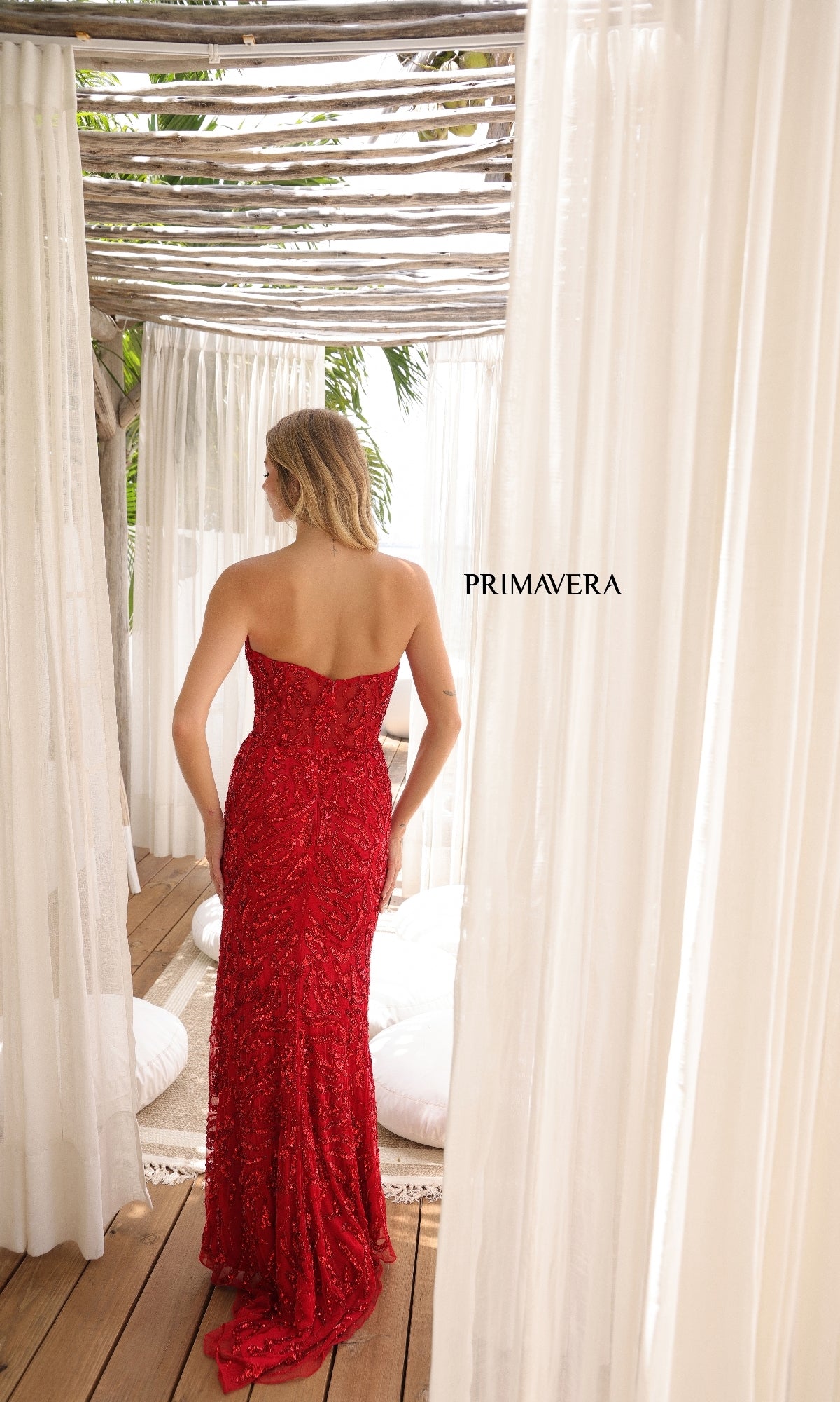 A woman wearing the Primavera 4360 long prom dress stands with her back to the camera between white curtains on a wooden floor, with 