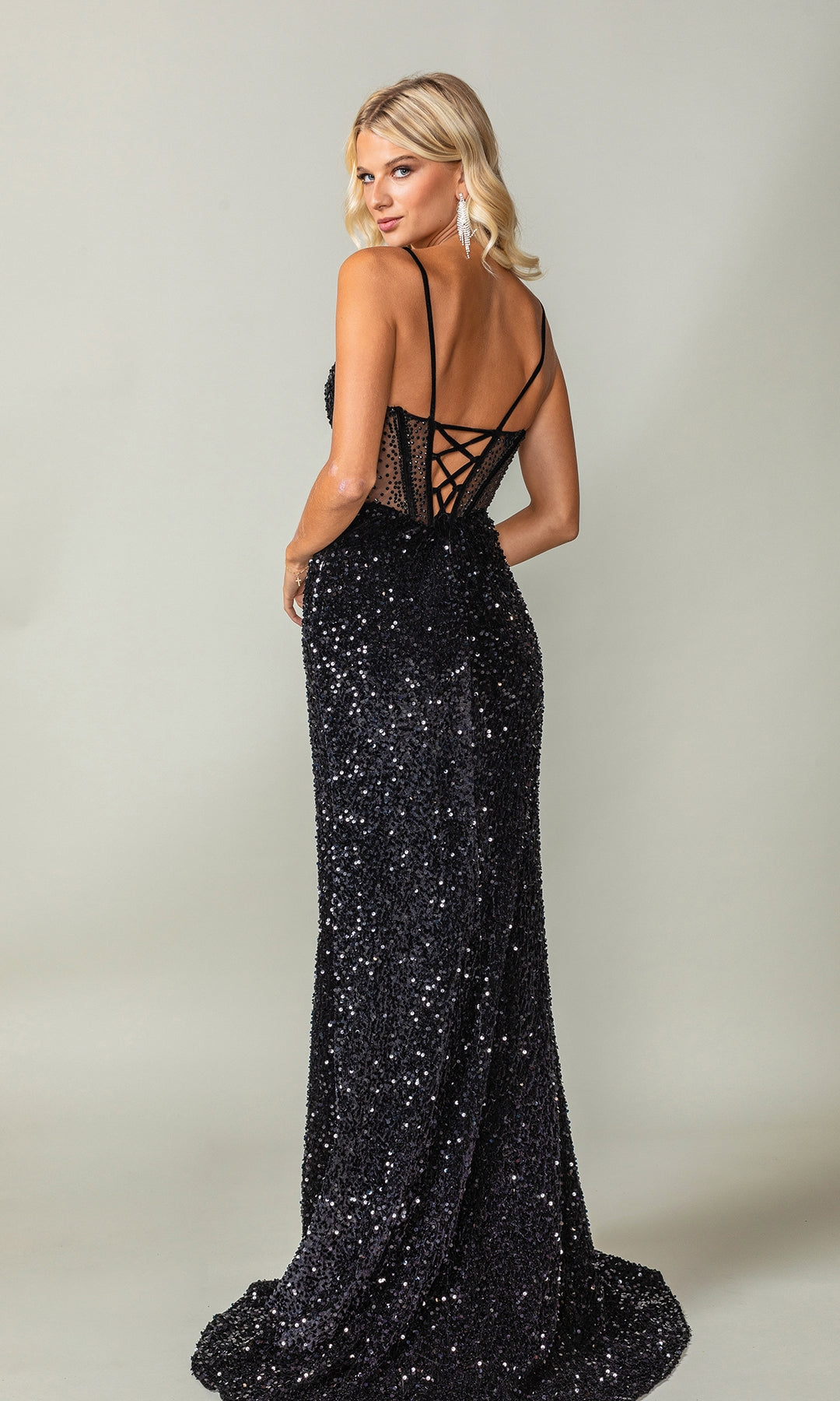 Woman wearing the Dancing Queen 4360: a floor-length, sleeveless black sequin gown with lace-up back, looking over her shoulder—an elegant prom dress perfect for making a statement.