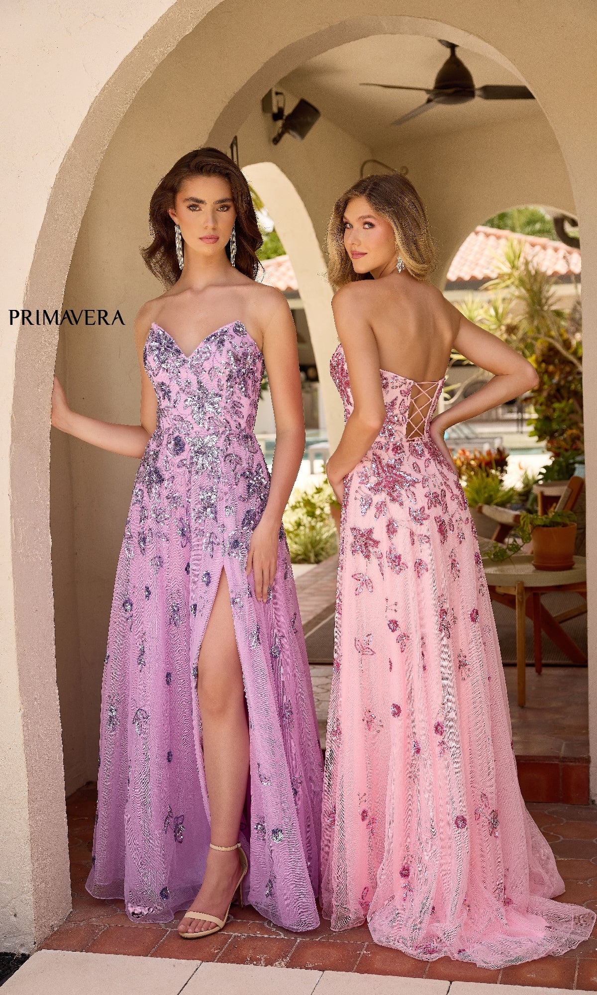 Two women in strapless embellished gowns—one in the lilac Strapless Beaded Long Prom Dress: Primavera 4361 with a thigh-high slit, the other pink with a lace-up back—stand by an outdoor archway decorated with patio furniture.