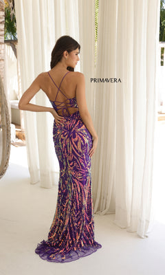 A woman faces away from the camera in the Strappy-Back Long Sequin Prom Gown: Primavera 4362, showcasing its crisscross open-back design with sparkling sequins. The word "PRIMAVERA" appears on the curtain beside her.