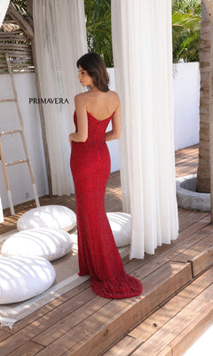 A woman wearing the Primavera 4364 long prom dress stands on a wooden deck, facing away, with white curtains, cushions, and a palm tree in the background.