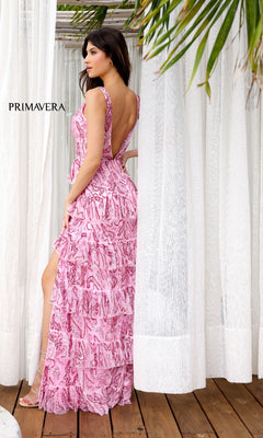 A woman stands on a wooden floor, surrounded by white curtains and tropical leaves, wearing the Primavera 4366 long prom dress—a pink, sleeveless gown with ruffles, a high slit, and tiered layers.