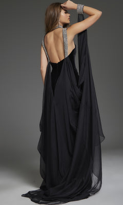 A woman stands facing away against a plain gray background, holding fabric over her arm while wearing the Jovani 43696 long black formal dress, featuring a beaded neckline and silver embellishments.