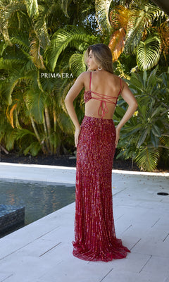 A woman in the Primavera 4370 Cut-Out Long Sequin Prom Dress, sparkling red, stands by a poolside patio with lush greenery in the background, facing away.