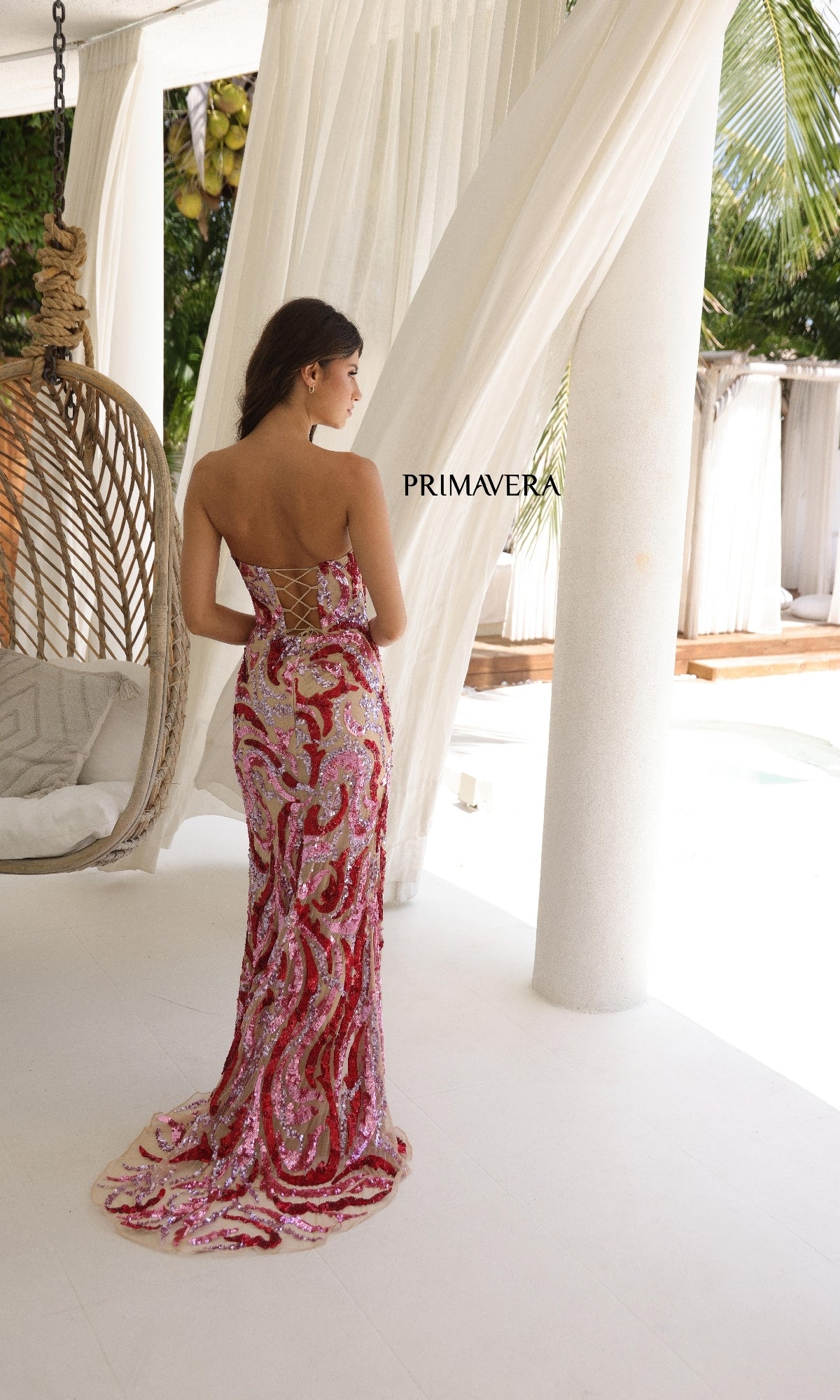 A woman wearing the Primavera 4373 Strapless Long Sequin Prom Dress in pink and red stands indoors by a hanging chair, facing away. The image features the word 