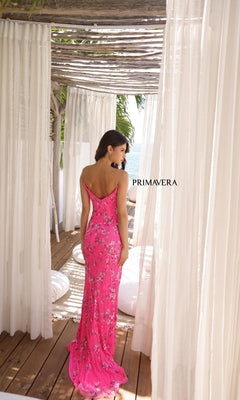 A woman wearing the Primavera 4374, a bright pink floral long prom dress, stands with her back to the camera on a wooden deck framed by sheer white curtains. "PRIMAVERA" appears beside her.