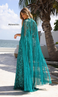 A woman stands outdoors near palm trees and water, facing away from the camera, wearing the Primavera 4376—a teal, sequined long prom dress with a sheer cape.