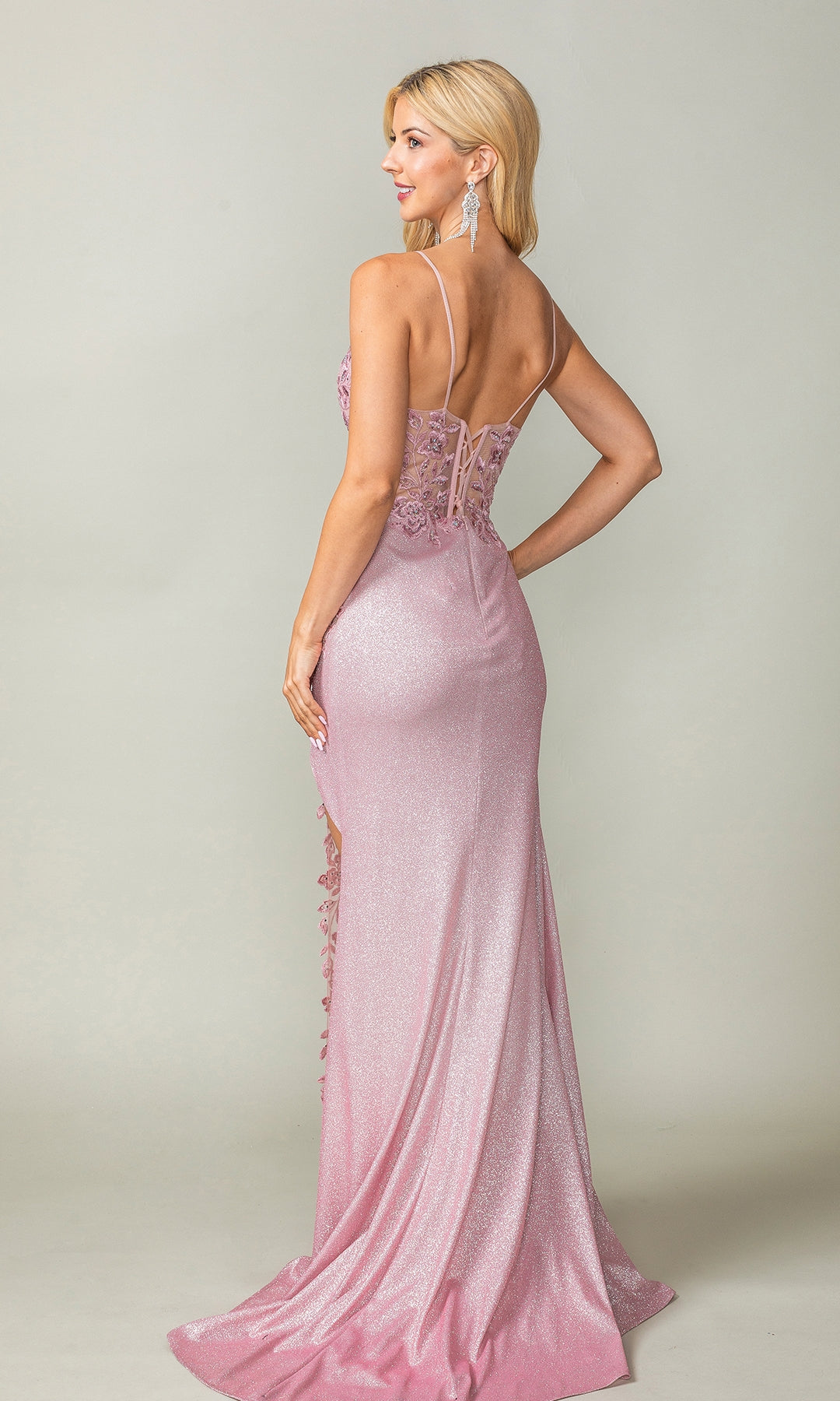 A woman poses against a plain light background wearing the Draped Long Glitter Prom Dress: Dancing Queen 4377—a shimmering light pink gown with thin straps, lace details, corset-style bodice, and low back.