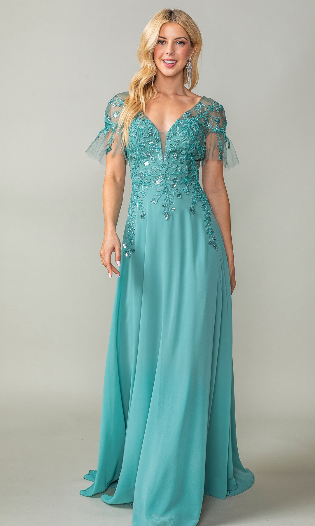 A woman poses against a plain light background in the Dancing Queen 4378 Sheer Short-Sleeve Prom Dress, featuring a floor-length teal design with floral sequin embroidery and a v-neckline.