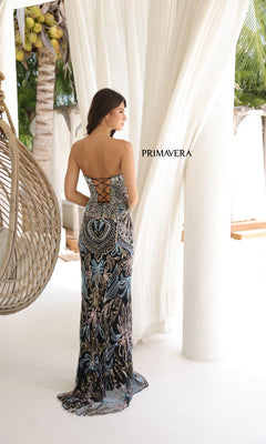 A woman wearing the Strapless Long Beaded Prom Dress: Primavera 4382, featuring a lace-up back, stands indoors near white curtains with tropical greenery visible outside.