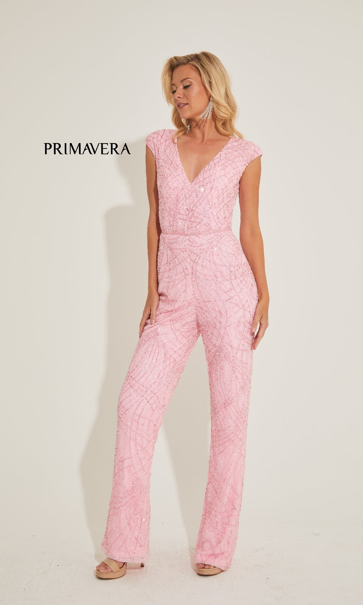 A woman stands against a plain background wearing the Primavera 4384 long prom jumpsuit, featuring light pink textured sequins, a V-neckline, and straight leg pants. 